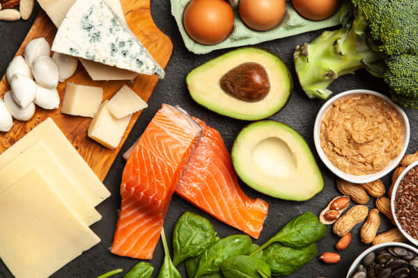 A variety of healthy foods including salmon fillets, sliced cheeses, eggs, avocado halves, broccoli, spinach, and nuts on a dark surface.