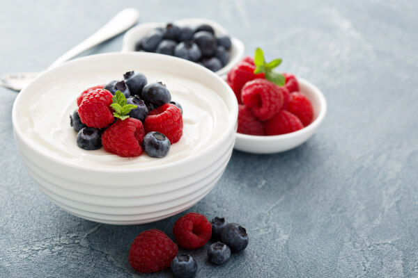 A white bowl of creamy yogurt topped with raspberries, blueberries, and mint, with bowls of more berries behind it on a gray countertop.
